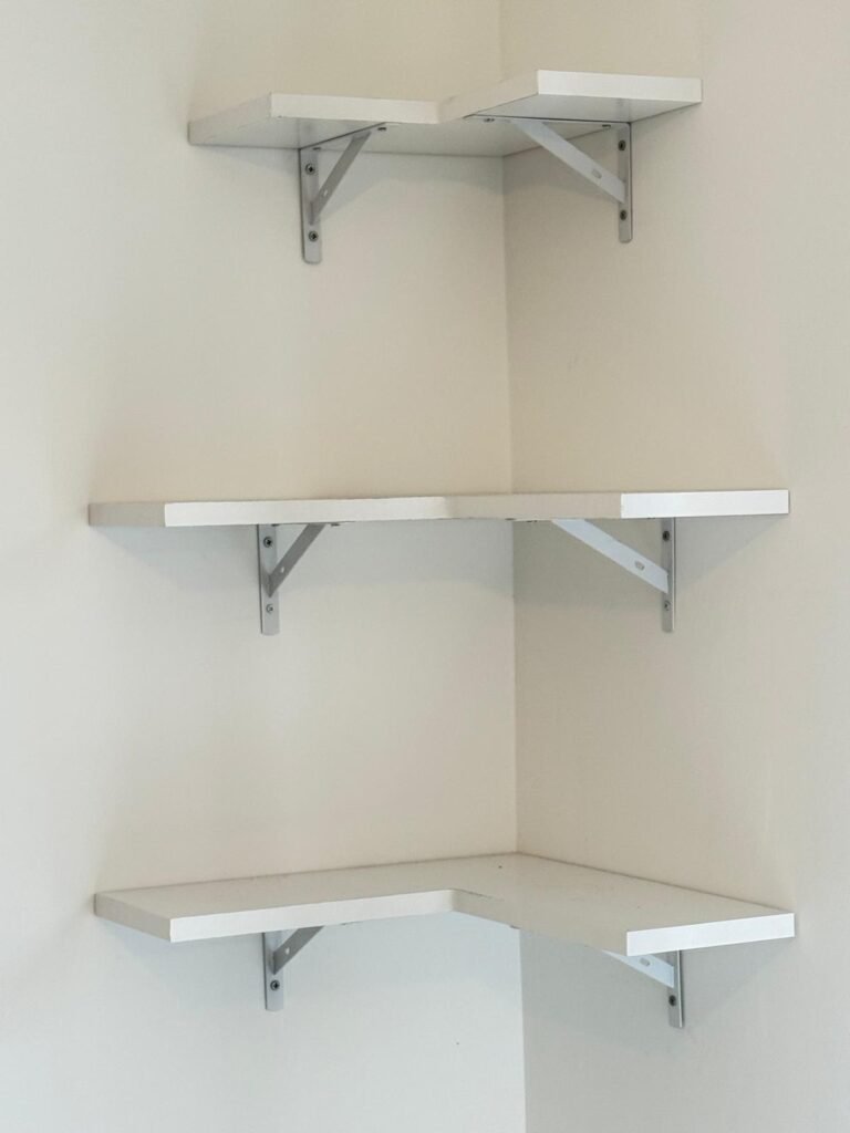 Three white corner shelves fitted level on brackets in a bedroom corner